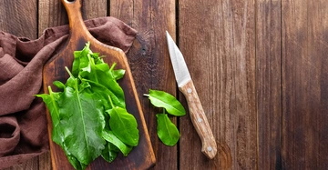 Frische grüne Blätter liegen auf einem Holzschneidebrett auf braunem Stoff eine Holzmesser liegt daneben alles auf einem rustikalen Holztisch.