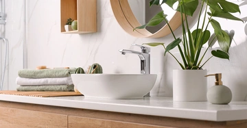 Waschbecken mit Wasserhahn steht auf einer weißen Ablagefläche Badezimmerpflanze und Seifenspender daneben an der Wand hängt ein Spiegel und Regal mit Dekoration im Hintergrund gestapelte Handtücher.