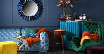 Blaues Sofa mit Kissen und Decke steht vor einer blauen Wand mit rundem Spiegel bunte Getränke und Blumenvasen auf Barhocker daneben grüner Sessel gelber Hocker und Teppich umgeben.