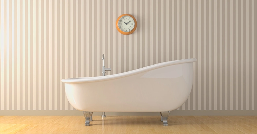 Eine weiße freistehende Badewanne steht auf einem hölzernen Boden vor einer beige gestreiften Wand. Eine runde Wanduhr hängt oberhalb der Badewanne.