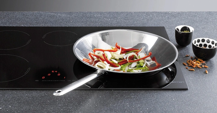 Eine große silberne Pfanne brät geschnittenes Gemüse auf einem modernen schwarzen Induktionskochfeld in einer Küche; daneben stehen zwei Schalen mit Gewürzen auf der Arbeitsplatte.