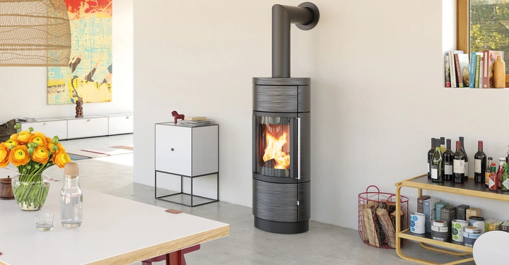 Ein moderner schwarzer Kaminofen mit brennendem Feuer steht vor einer weißen Wand in einem Wohnzimmer. Daneben befinden sich ein Regal mit Weinflaschen und Vorräten sowie ein Tisch mit einem Blumenstrauß.