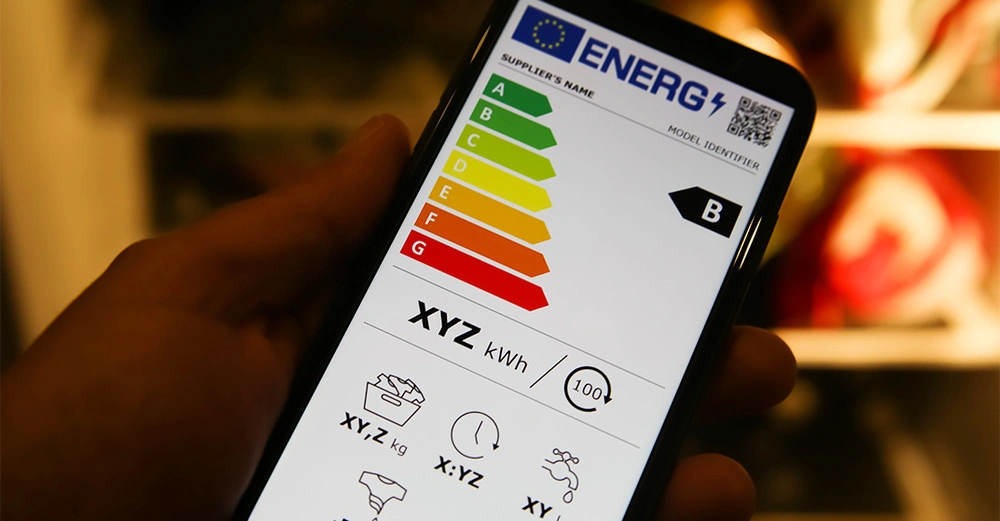 Eine Hand hält ein Smartphone das ein Energielabel mit einer Skala von A bis G zeigt wobei B markiert ist. Text vorhanden Energ XYZ kWh.