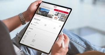 Person hält ein Tablet zeigt eine Immobilienanzeige mit Preis 990000 € Standort 2831 Wiesenhof Fläche 285 m² 10 Zimmer und weitere Details; Umgebung: unscharfes Wohnzimmer.