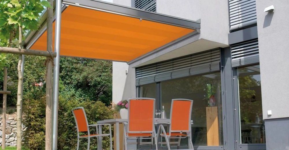 Ein Terrassentisch mit vier Stühlen steht unter einer ausfahrbaren orangefarbenen Markise vor einem modernen Haus mit großen Glasfenstern. In der Umgebung gibt es Grünflächen und Büsche.