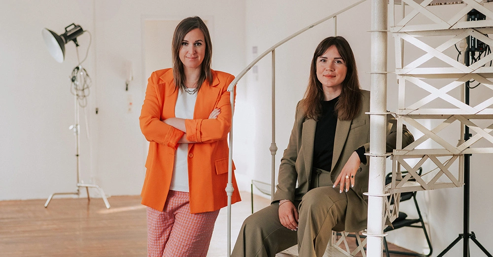 Zwei Frauen mit dunklen, schulterlangen Haaren. Links in orangen Hosenanzug gekleidet und stehend mit verschränkten Armren. Rechts auf weißer Wendeltreppe sitzend, in graubraunem Hosenanzug. 