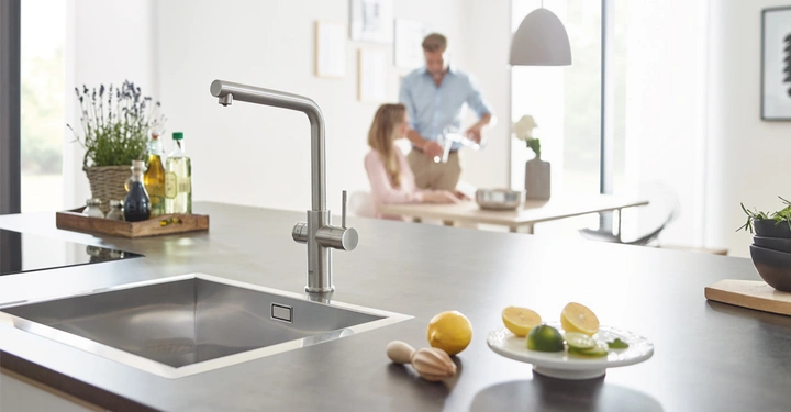 Edelstahl-Wasserhahn stillstehend in einer modernen Küche mit Kräutern und Zitronen auf der Arbeitsfläche im Vordergrund; im Hintergrund gießen zwei Personen Wasser ein und sitzen am Esstisch.