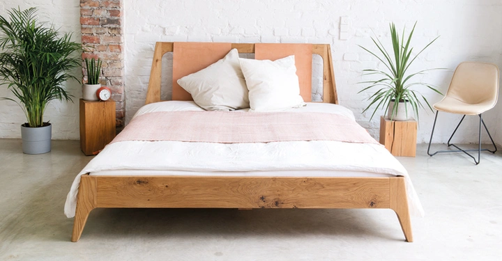 Holzbett mit weißen Kissen und rosafarbener Decke steht in einem minimalistischen Schlafzimmer mit weißen Wänden Pflanzen und einem Stuhl im Hintergrund