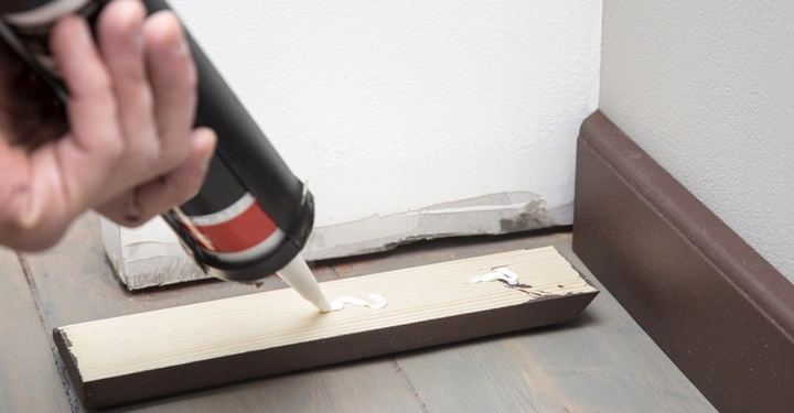 Hand trägt Klebstoff aus einer Kartusche auf ein Holzstück auf, das auf einem Holzboden in der Ecke eines Raums mit einer beschädigten Wand und dunkelbraunem Sockelbrett liegt