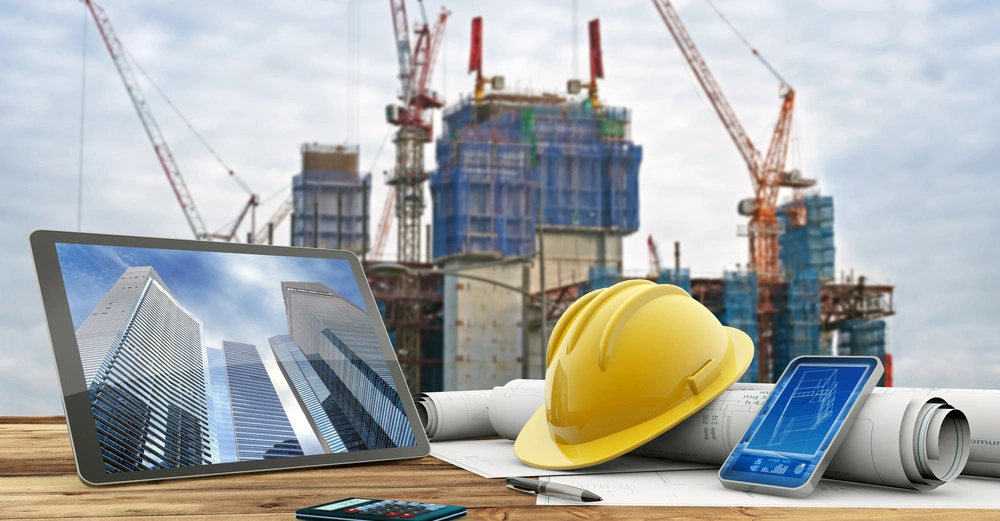 Tablet und gelber Bauhelm auf Zeichnungen liegend vor Baustelle mit Kränen und Hochhäusern im Hintergrund