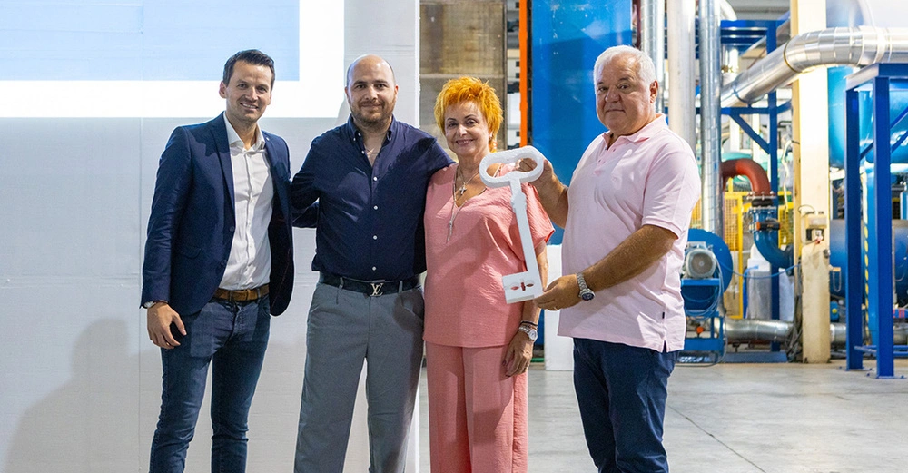 3 Männer und eine Frau stehen nebeneinander in Fertigungshalle und lächeln in die Kamera. Symbolische Schlüsselübergabe für Expansionsabschluss. 