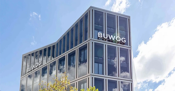 Hochhaus mit dem Schriftzug BUWOG an der Glasfassade baum im Vordergrund und Himmel mit wenigen Wolken im Hintergrund.