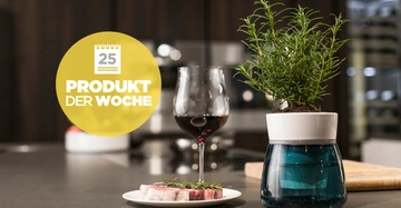 Pflanze steht neben einem Glas Rotwein und einem Teller mit Fleischstück in einer modernen Küche Text im gelben Kreis 25 PRODUKT DER WOCHE.