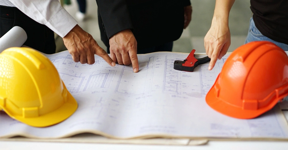 Drei Personen stehen vor einem Tisch mit Bauplänen und deuten auf die Unterlagen. Im Vordergrund liegt ein gelber und ein orangefarbener Schutzhelm.
