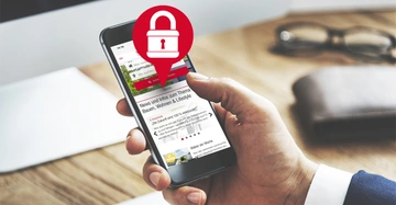 Eine Hand hält ein Smartphone welches eine Nachrichten-Website zeigt. Ein rotes Schloss-Symbol über dem Bildschirm deutet auf Sicherheit hin. Im Hintergrund ein Schreibtisch mit Brille und Notizbuch.