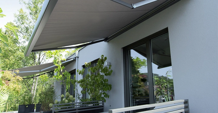 Eine große Markise erstreckt sich über eine moderne Terrasse mit Holzboden, umgeben von Pflanzen und Bäumen, neben einem weißen Gebäude mit großen Fenstern und grauen Gartenbänken.