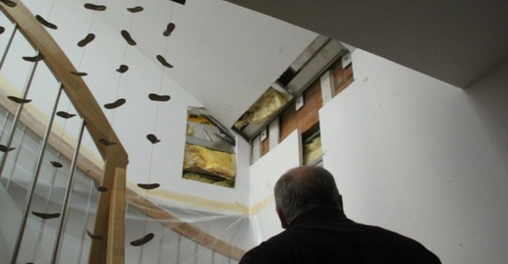 Galerie-Bild 29: Ein Mann schaut eine teilweise entkernte Wand in einem modernen Treppenhaus an die Wand zeigt isolierte Bereiche und freiliegende Baumaterialien eine dekorative Holztreppe ist im Vordergrund zu sehen.