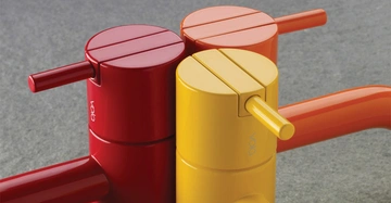 Drei bunte Wasserhähne in Rot Gelb und Orange befinden sich nebeneinander auf einer grauen Oberfläche. Die Wörter Vola sind auf den gelben und roten Wasserhähnen sichtbar.