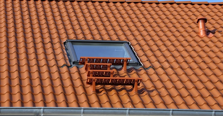 Dachfenster eingebaut in ein orangefarbenes Ziegeldach mit Dachtrittstufen und Lüftungsschlot in einem sonnigen Außenbereich.