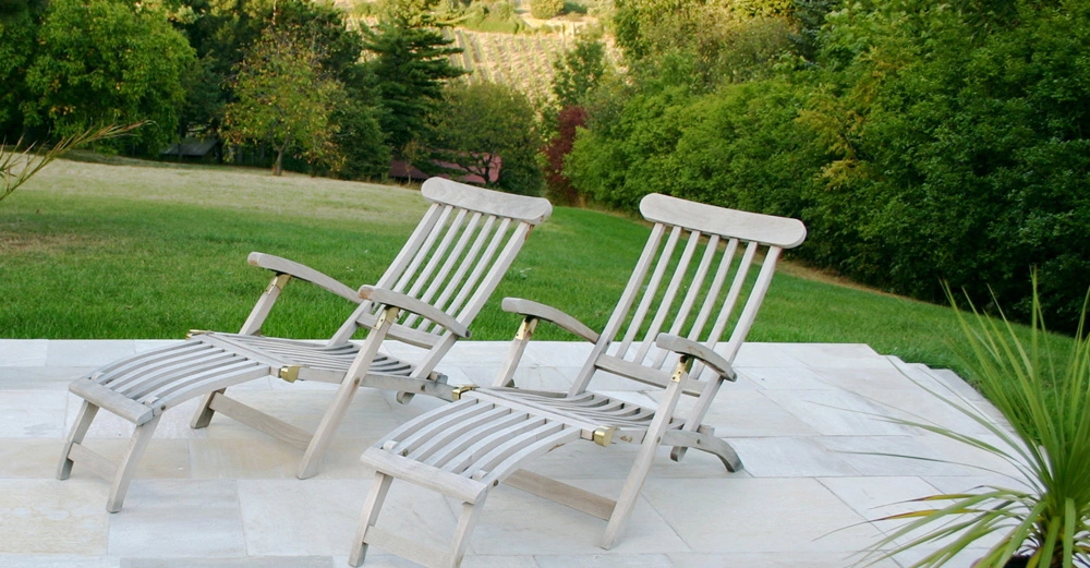Zwei Holzliegen stehen nebeneinander auf einer Steinplatte. Im Hintergrund erstreckt sich ein grüner Garten mit Bäumen und Hügeln. Links eine große Grünfläche, rechts eine Topfpflanze.