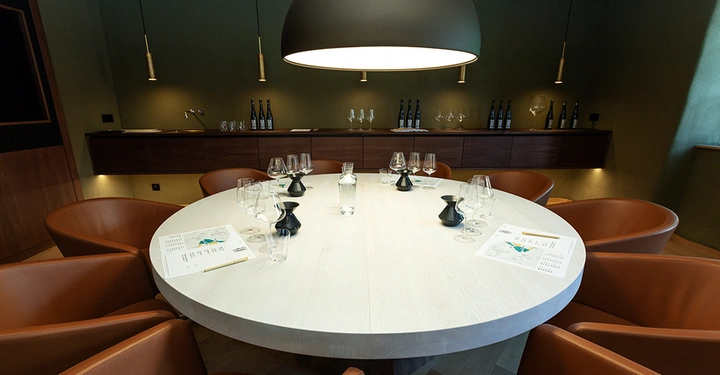 Galerie-Bild 10: Ein großer runder Tisch ist mit Weingläsern und Karaffen gedeckt und von braunen Ledersesseln umgeben. Im Hintergrund stehen Weinflaschen und Gläser auf einem Regal unter einer modernen Hängelampe.