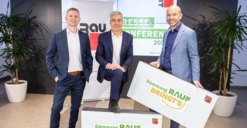 Drei Männer posieren vor einer Wand mit einem Poster in einem Raum mit Pflanzen der mittlere Mann hat ein Bein auf eine Stufe gestellt und die beiden anderen halten ein Schild. Text im Bild Rau Pressekonferenz 202 Dämmung RAUF BRINGT'S! Baumit. Ideen mit Zukunft.