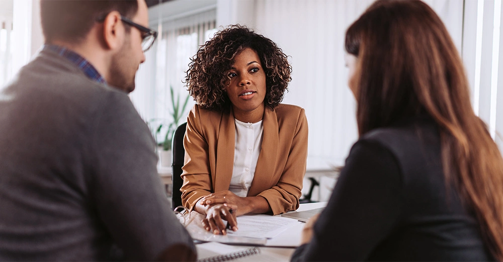 Drei Personen sitzen an einem Tisch und besprechen Dokumente im Büro mit Fenstern im Hintergrund