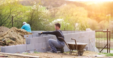 Arbeiter setzt Betonblöcke auf eine Mauer in einem offenen, grünen Bereich, während im Hintergrund ein weiterer Arbeiter mit Erde arbeitet. Eine Schubkarre steht neben dem Arbeiter.