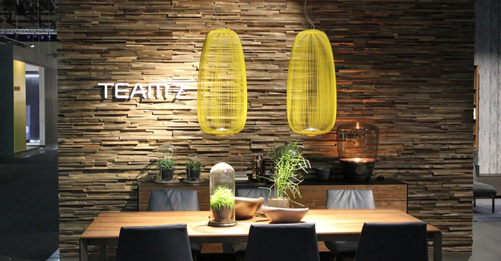 Galerie-Bild 14: Holztisch mit Schalen und Pflanzen darauf steht vor einer Wand mit aufgesetzten Holzpanele; zwei gelbe Lampen hängen darüber. Wandtext: TEAM 7. Umgebung: modern eingerichtet.