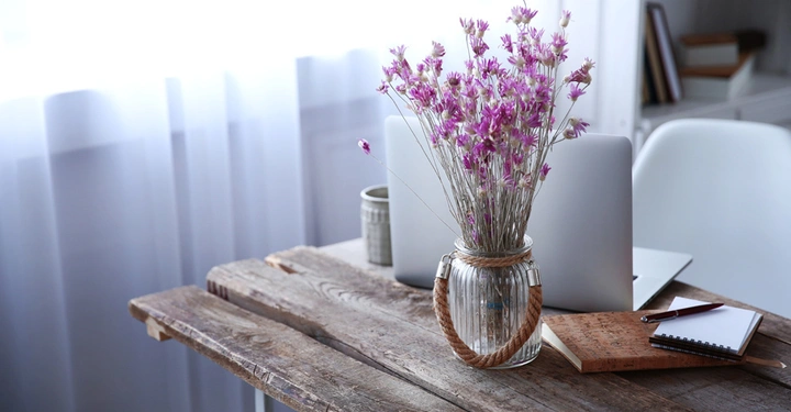 Ein Glasvase mit rosa Blumen steht auf einem rustikalen Holztisch zusammen mit einem Laptop einer Kaffeetasse Notizbüchern und einem Stift in einem hellen und gemütlichen Arbeitszimmer.