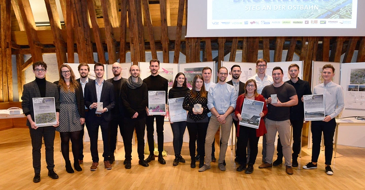 Menschen halten Poster und Trophäen Präsentation in einem Holzraum Plakate und ein Projektionsbildschirm zeigen Brückenbau Steg an der Ostbahn. Text: BRÜCKENBAU STEG AN DER OSTBAHN