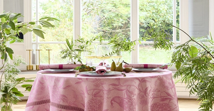 Runder Esstisch elegant gedeckt mit einer rosaroten Leinentischdecke mit Pfauen darauf. Auf dem Tisch stehen Platzteller, auf ihnen liegen in einem goldenen Serviettenring die passenden Stoffservietten.