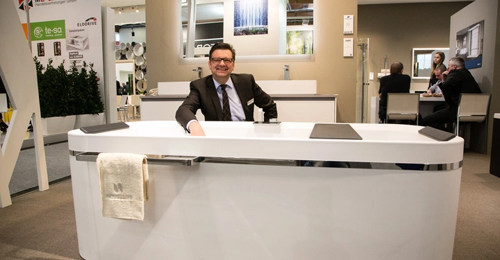 Galerie-Bild 4: Ein Mann sitzt hinter einem weißen Schreibtisch und lächelt in einer Messeumgebung mit mehreren Ständen im Hintergrund. Auf dem Schreibtisch liegt ein Handtuch mit der Aufschrift NOVELLINI. Ein Schild zeigt: ME-GA Handelsvertretungen GmbH te-sa ELODRIVE.