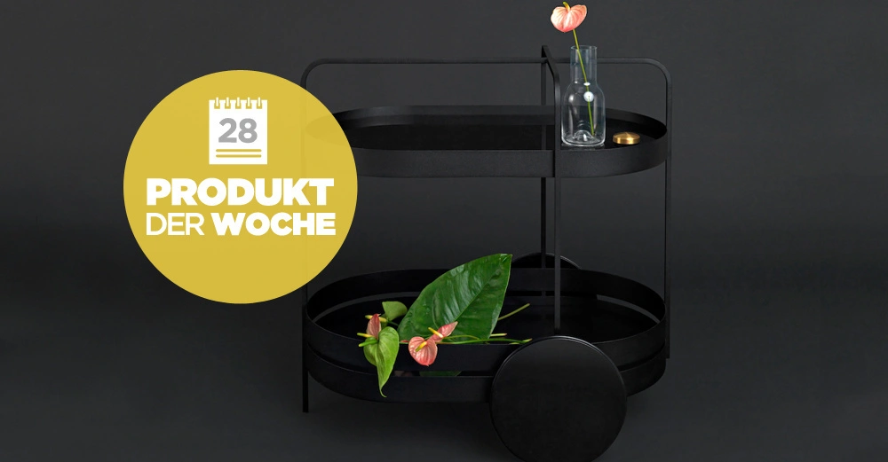 Schwarzer Servierwagen hält oben eine Vase mit Blume und unten Blätter auf dunklem Hintergrund. Gelbes Symbol zeigt Text ''28 PRODUKT DER WOCHE''.