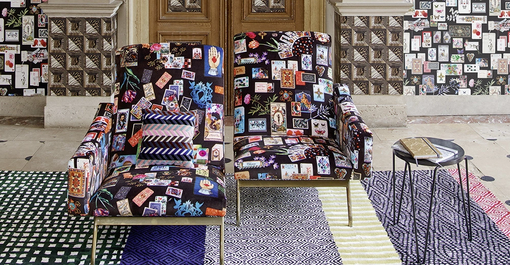Zwei bunte Sessel mit Aufdruck von Karten und Blumenmuster stehen nebeneinander in einem Raum; ein kleiner Beistelltisch steht rechts daneben auf einem gemusterten Teppich vor einer Wand mit ähnlichem Muster.