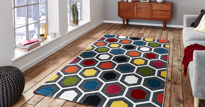 Galerie-Bild 4: Ein bunter Teppich mit sechseckigen Mustern liegt auf einem Holzboden neben einem grauen Sofa. Im Hintergrund stehen ein hölzernes Sideboard und große Fenster mit Stadtblick. Text: INSPIRATION, CONRAN.