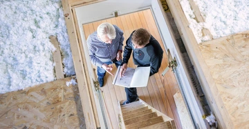 Zwei Männer besprechen Dokumente eines Klemmbretts in einem Raum mit Holzfußboden auf einer Treppe. Die Umgebung scheint eine Baustelle mit Isolationsmaterial zu sein.
