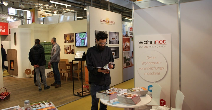 Galerie-Bild 19: Ein Mann blättert durch ein Magazin an einem Wohnnet-Stand in einem Ausstellungsraum mit mehreren Ständen. Text auf Plakat: Deine Wohntraumverwirklichungsmaschine. Andere Leute schauen sich Exponate an.