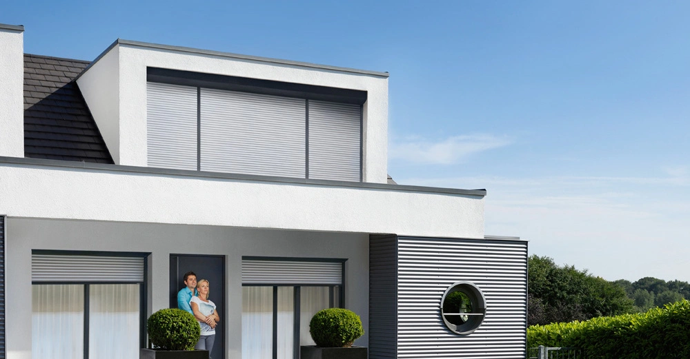 modernes Einfamilienhaus mit grüner Hecke und geschotterter Einfahrt. Paar steht Arm in Arm vor Eingangstür, große Fensterflächen mit halb heruntergelassenen Rollläden in hellgrau von Schlotterer