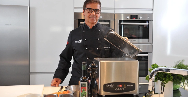 Ein Koch in schwarzer Uniform öffnet ein Sous-vide-Gerät in einer modernen Küche. Auf der weißen Arbeitsplatte befinden sich Gewürze, Öl und frisches Gemüse. Text: Iavide Beste Qualität.
