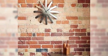 An der Ziegelwand hängt eine moderne Uhr mit strahlenförmigen Metallzeigern über einem Holztisch auf dem zwei braune Flaschen stehen