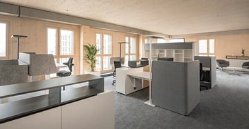 Büromöbel stehen geordnet in einem offenen Raum Büroarbeitsplätze mit Trennwänden und Schreibtischen mehrere Bürostühle nahe großer Fenster Holzverkleidung an den Wänden und ein Teppichboden einige Pflanzen zur Dekoration