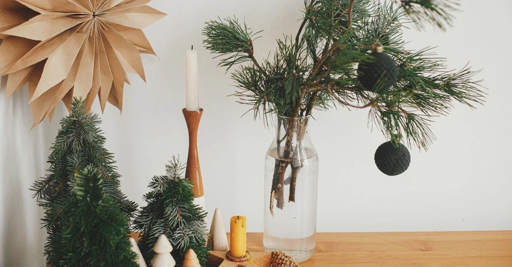 Mehrere Weihnachtsdekorationen wie miniatur Weihnachtsbäume stehen auf einem Holztisch zusammen mit einem Glasgefäß mit Tannenzweigen und einem Papierstern an der Wand im Hintergrund.
