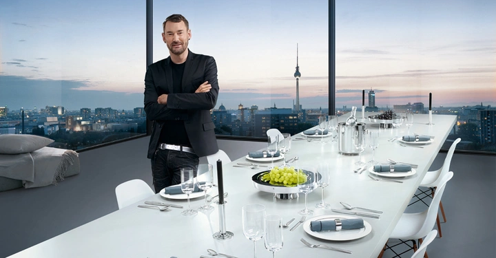 Ein Mann steht mit verschränkten Armen am Kopfende eines langen, festlich gedeckten Esstisches vor einer Glaswand, die einen Blick auf eine Stadtlandschaft bei Sonnenuntergang bietet