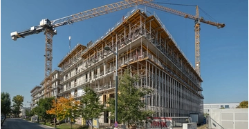 Der Bauprozess der Zentralberufsschule Seestadt Aspern in Wien mit Gerüst und Kränen vor einem blauen Himmel.