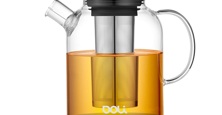 Ein transparenter Glasteekessel mit Metallfilter in der Mitte und einem schwarzen Griff steht auf einer weißen Oberfläche. Wasser ist orange gefärbt. Text Doli auf der Kanne.