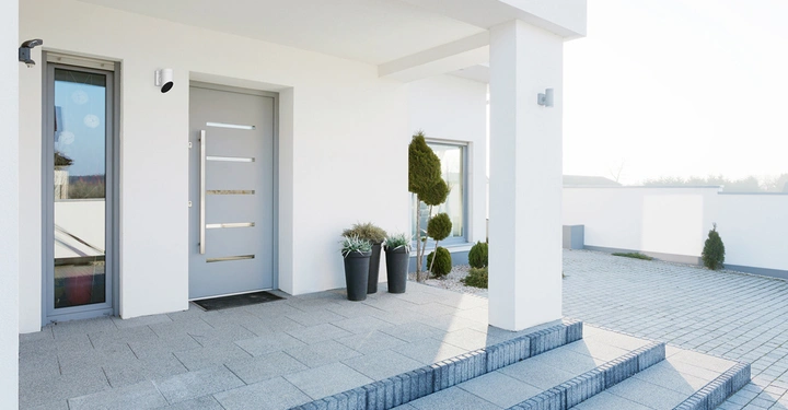 Graue Haustür bleibt still weiße Gebäudefassade umgeben von Steinfliesen drei Stufen Eingang vorgesetzte Pflanzen in Töpfen Sichtkamera an der Wand großer Innenhof im Hintergrund