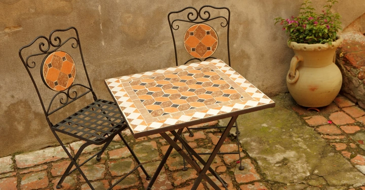 Galerie-Bild 6: Sitzecke auf einer rustikalen toskanischen Terrasse. Auf dem altem Steinboden stehen ein eckiger Mosaiktisch in Erdtönen und zwei schwarze Metallstühle mit runder Rückenlehne aus Mosaik.