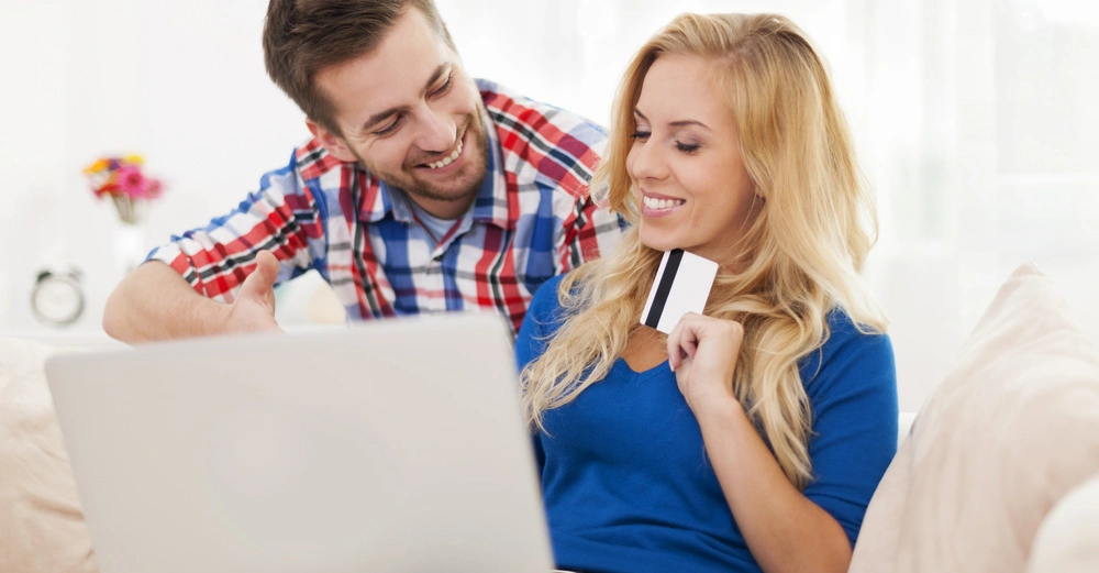 Ein Mann zeigt auf einen Laptop und eine Frau hält eine Kreditkarte in der Hand beide lächelnd auf einem Sofa sitzend im Wohnbereich mit unscharfer Dekoration im Hintergrund.
