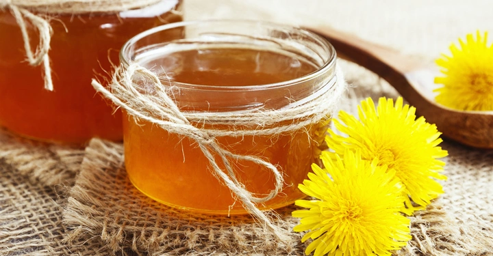 Ein Glas mit bernsteinfarbenem Honig dekoriert mit Jutegarn steht auf einem Jutestoff links daneben ein weiteres Glas und gelbe Löwenzahnblüten im Hintergrund ein Holzlöffel mit einer Blüte